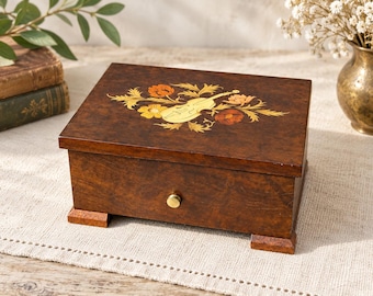 Puede incluir: Una caja de música rectangular de madera marrón oscuro con acabado pulido y cuatro patas pequeñas. La tapa presenta un diseño de marquetería incrustada de un violín rodeado de flores naranjas, amarillas y rojas. Un pequeño pomo de latón está centrado.