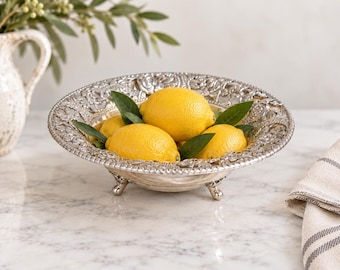 Puede incluir: Un cuenco de metal plateado con un borde floral en relieve y tres patas pequeñas. El cuenco está lleno de cuatro limones de color amarillo brillante y varias hojas verdes, mostrando un acabado metálico reflectante y detalles decorativos.