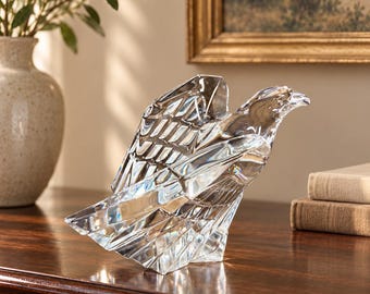Puede incluir: Una escultura de cristal transparente de un águila en posición posada con las alas parcialmente plegadas. La figura presenta superficies facetadas que captan la luz, creando reflejos geométricos nítidos en el cuerpo, la cabeza y la cola.