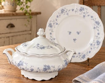 Puede incluir: Una sopera de cerámica blanca con tapa y plato a juego, ambos decorados con un delicado patrón floral azul. La sopera cuenta con dos asas laterales y una base festoneada, mientras que el plato presenta un borde festoneado y un motivo central.