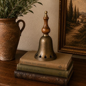 May include: A brass bell with a wooden handle sits atop a stack of three books. The bell features a flared base with cut-out shapes. The books are titled Trees and Shrubs and Garden Flowers. The bell has a metallic finish and a dark wood top.
