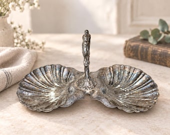 Puede incluir: Un plato doble de metal plateado que presenta dos cuencos unidos en forma de concha. Un asa vertical central se eleva desde el medio, rematada con una pequeña figura esculpida. La superficie metálica muestra una pátina desgastada y oscura.