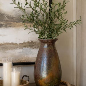 May include: A tall, pear-shaped ceramic vase with a mottled, iridescent finish in shades of deep brown, bronze, and green. The vase contains a cluster of green leafy branches. Beside it sits a woven tray with two white pillar candles.