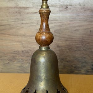 May include: A brass bell featuring a turned wooden handle with a rounded center and a small brass finial at the top. The bell body is made of aged brass and includes a row of small, evenly spaced cutouts shaped like apples along the bottom edge.