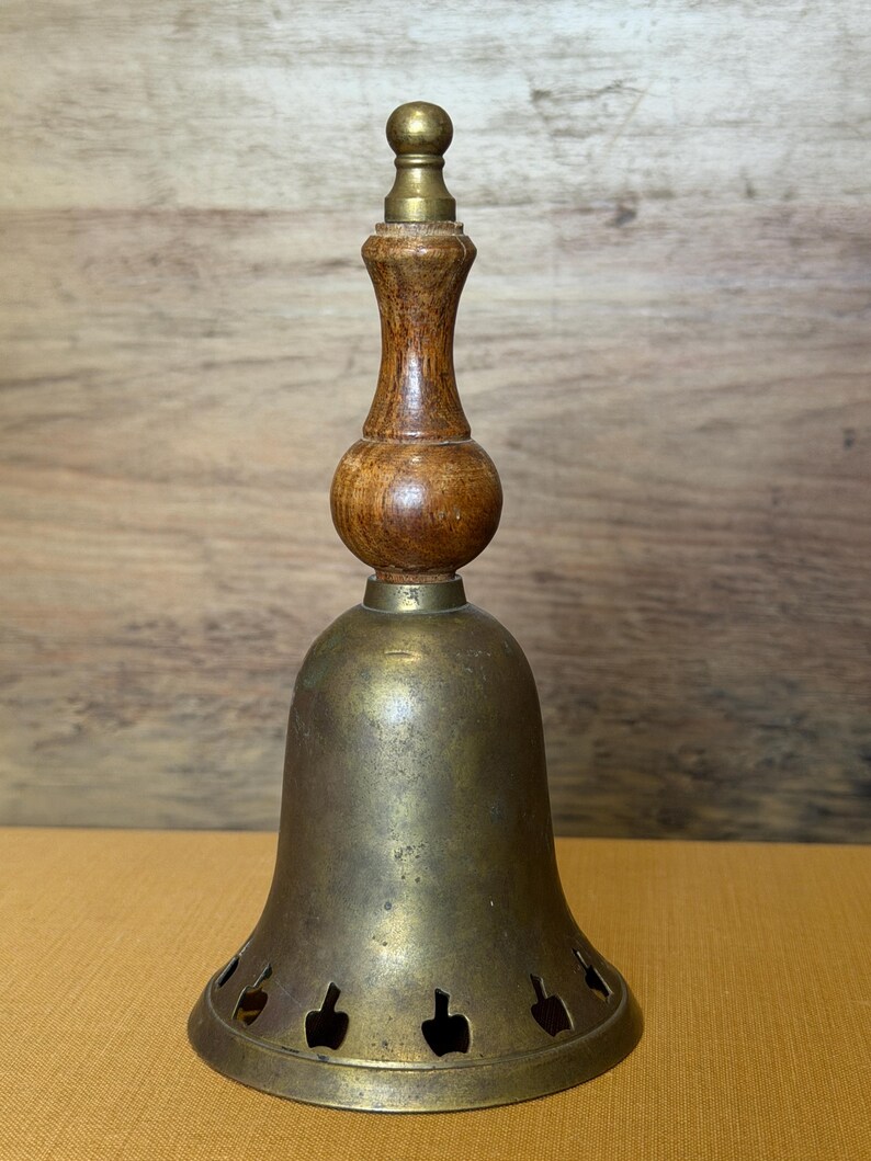 May include: A brass bell featuring a turned wooden handle with a rounded center and a small brass finial at the top. The bell body is made of aged brass and includes a row of small, evenly spaced cutouts shaped like apples along the bottom edge.