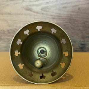 May include: A brass bell with a circular opening featuring a series of cut-out shapes along the rim. Inside the bell, a metal clapper hangs from a small loop, positioned to strike the interior wall to produce sound when the bell is moved.