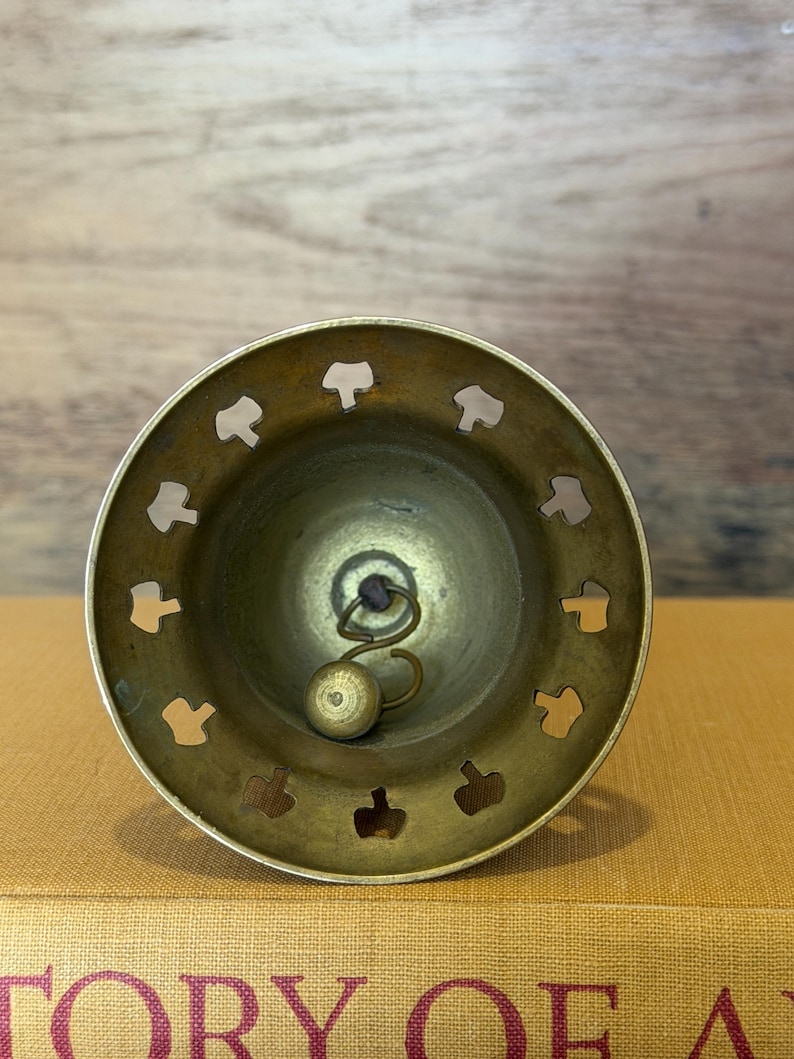 May include: A brass bell with a circular opening featuring a series of cut-out shapes along the rim. Inside the bell, a metal clapper hangs from a small loop, positioned to strike the interior wall to produce sound when the bell is moved.