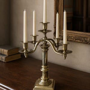Puede incluir: Un candelabro de latón con un pilar central y cuatro brazos curvos, cada uno con una vela blanca delgada. El metal tiene un acabado pulido con signos visibles de antigüedad y pátina, una base cuadrada y elementos decorativos escalonados.