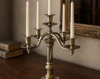 May include: A brass candelabra with a central pillar and four curved arms, each holding a slender white taper candle. The metal has a polished finish with visible signs of age and patina, featuring a square base and tiered decorative elements.