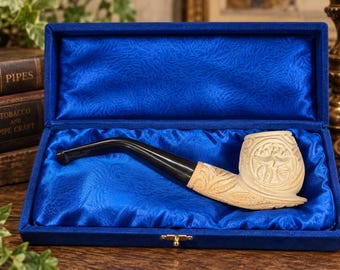 Puede incluir: Una pipa de tabaco con un hornillo tallado de color crema que presenta intrincados patrones de hojas y círculos, unida a una boquilla negra lisa. La pipa se presenta dentro de un estuche rectangular forrado con tela de satén azul.