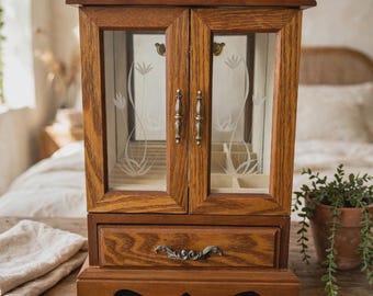 Puede incluir: Un joyero de madera con dos puertas de cristal que presentan diseños florales grabados y herrajes en tono latón. El mueble incluye un único cajón inferior con un tirador de metal ornamentado y un compartimento interior con varias ranuras.