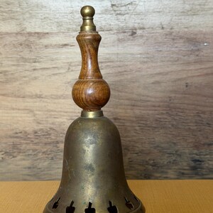 May include: A hand bell featuring a tarnished brass body with a series of small, cut-out shapes along the bottom edge. The handle is crafted from polished brown wood with a smooth, rounded center section and a small brass finial at the very top.