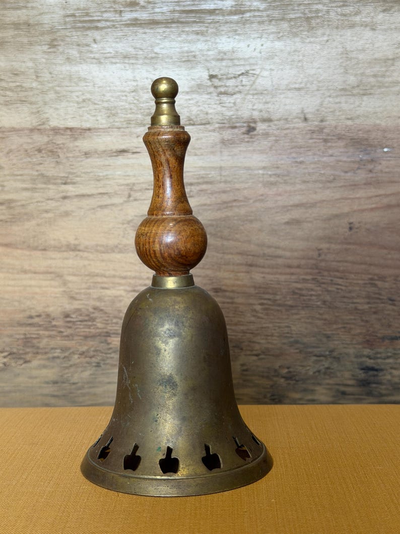 May include: A hand bell featuring a tarnished brass body with a series of small, cut-out shapes along the bottom edge. The handle is crafted from polished brown wood with a smooth, rounded center section and a small brass finial at the very top.