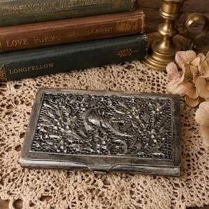 May include: A rectangular silver-toned metal case featuring an intricate, raised relief design of a bird surrounded by dense floral and foliage patterns. The surface has a dark, oxidized finish within the recessed areas, highlighting the detailed metalwork.