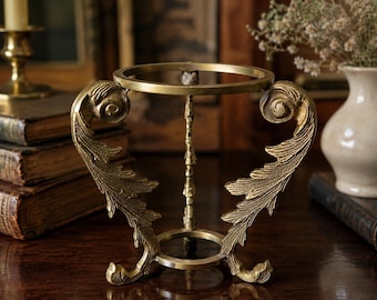May include: A gold-toned metal display stand featuring a circular top ring and a circular base connected by three ornate, curved supports. Each support displays detailed leaf-like carvings and scrollwork, creating a structured frame for holding objects.