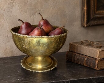 Puede incluir: Un cuenco de pedestal en tono dorado metálico con acabado texturizado y borde festoneado, que contiene cuatro peras de color rojo oscuro. El cuenco presenta una base circular ancha y se sitúa junto a dos libros antiguos con gafas.