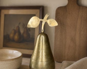 May include: A pear-shaped decorative object crafted from polished brass. The item features a smooth, rounded body and a distinct stem at the top with two flat, leaf-like protrusions extending horizontally from the center of the stem.