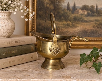 Puede incluir: Un cubo para carbón de latón pulido con un cuenco redondeado, una base de pedestal y un asa vertical. El recipiente está adornado con un emblema decorativo de cabeza de león en la base del asa e incluye un asa lateral horizontal.