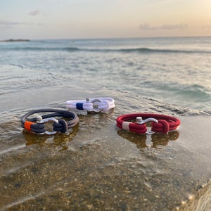 May include: Three nautical-style bracelets with stainless steel clasps. One is dark gray with an orange accent, another is white with blue, and the third is red with a beige accent. The bracelets are displayed on a wet, stone surface with the ocean in the background.