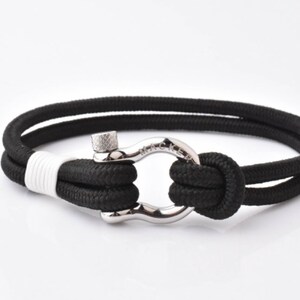 Pode incluir: Pulseira de corda preta com fecho de metal prateado. O fecho é um laço com um pequeno botão de metal na parte superior.