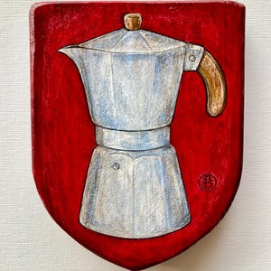 May include: A painted shield-shaped plaque featuring a silver moka pot with a brown handle centered on a vibrant red background. The artwork displays a textured finish with visible brushstrokes and a small circular red stamp in the lower right corner.