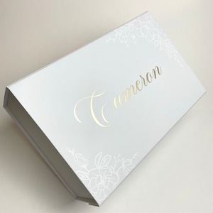 May include: A white gift box with the name "Cameron" in gold script. Floral designs in white are in the corners. The box is rectangular and has a magnetic closure.
