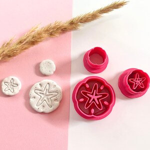 May include: A set of four pink plastic cookie cutters shaped like sand dollars and starfish. The cutters are on a white surface with a dried grass stem.