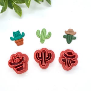 May include: Set of six red plastic cookie cutters shaped like cacti. The cutters feature different cactus designs, including a cactus with a cowboy hat, a cactus with a cat face, and a traditional cactus.