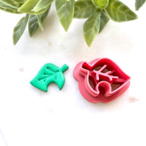 May include: A green leaf-shaped item and a pink leaf-shaped cookie cutter are displayed on a white surface. The green leaf has detailed veins. The pink cutter has a similar leaf design with a rounded edge. Green foliage is in the background.