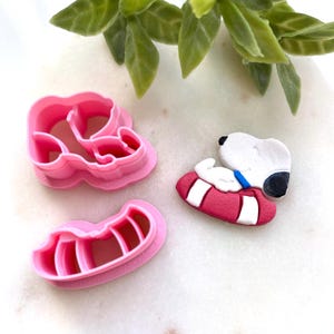 May include: Pink plastic cookie cutters in the shape of Snoopy and his doghouse. A Snoopy figurine is also present, lying on a red and white striped cushion. The items are on a white surface with green foliage.