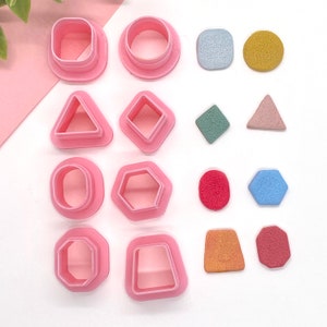 May include: A set of 10 pink plastic cookie cutters in various shapes, including a circle, square, triangle, diamond, hexagon, and a rounded rectangle. The cutters are arranged on a white surface with colorful decorative accents.