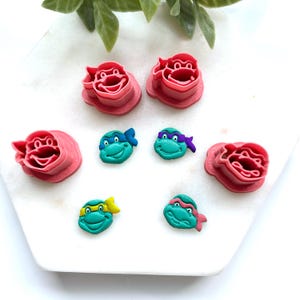 May include: A collection of Teenage Mutant Ninja Turtle-themed polymer clay charms and cookie cutters. The charms feature the turtles' faces with colored masks. The cookie cutters are pink and shaped like the turtles' faces. The items are on a white surface.