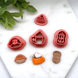May include: A selection of pink plastic cookie cutters in various shapes, including a coffee cup, a peanut, and a pie. There are also several finished clay pieces, including a coffee cup with whipped cream and a peanut.