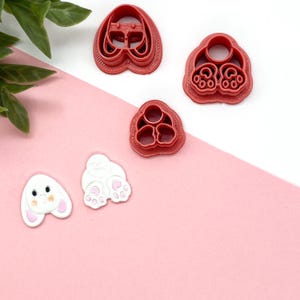 May include: Four red plastic cookie cutters shaped like a bunny rabbit. The cutters are on a white and pink surface. The cutters are for making cookies, fondant, or other edible crafts.