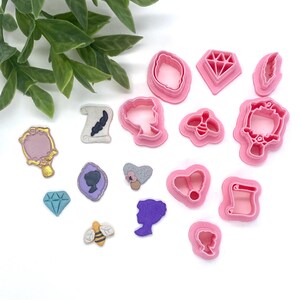 May include: A set of 14 pink plastic cookie cutters in various shapes, including a bee, a heart, a diamond, a scroll, a mirror, a crown, a flower, a silhouette of a woman, and a silhouette of a woman's head.