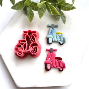 May include: A collection of scooter-themed items. A pink scooter cookie cutter is next to two scooter-shaped embellishments, one light blue and one pink. The scooters have black tires and are on a white surface with green leaves.