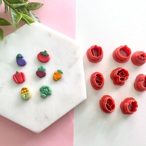May include: A set of 10 red plastic cookie cutters shaped like various vegetables, including a carrot, eggplant, tomato, and broccoli. The cutters are arranged on a white surface.