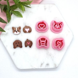 May include: Six pink plastic cookie cutters shaped like dogs and hearts. The cutters are on a white marble surface.