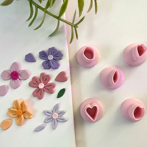 May include: A set of pink polymer clay flower and heart-shaped molds. The molds are arranged on a white surface. The molds are used to create polymer clay flowers and hearts.