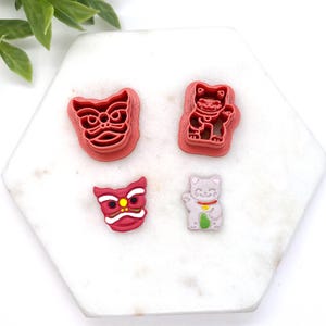 May include: Four red and white cookie cutters shaped like a lion, a cat, and two different versions of a dragon. The cookie cutters are on a white marble surface.