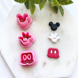 May include: Pink plastic cookie cutters in the shape of Mickey Mouse's head, gloves, and shorts. Also, black Mickey Mouse head, white glove, and red shorts decorations. The items are arranged on a white marble surface.