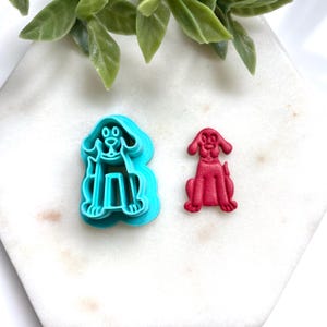 May include: A bright turquoise plastic dog-shaped cookie cutter alongside a matching red dog-shaped cutout. The cutter features a detailed outline of a seated dog with floppy ears, while the red cutout displays the same shape in a solid form.