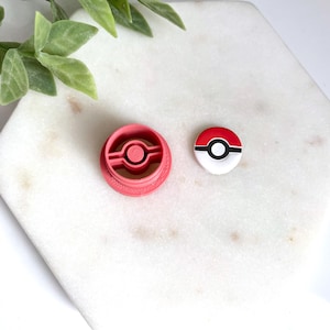 May include: A pink plastic cookie cutter and a round button with a red, white, and black design. The cookie cutter is circular with a central circle. The button has a black stripe separating the red and white sections. The items are on a white surface.