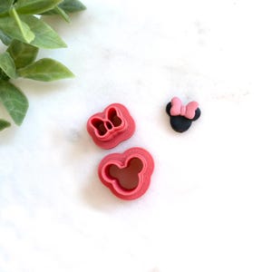 May include: Three items are displayed on a white surface: two pink cookie cutters in the shape of a bow and a mouse head, and a small black mouse head with a pink bow. The cookie cutters are made of plastic.