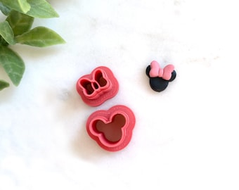 May include: Three items are displayed on a white surface: two pink cookie cutters in the shape of a bow and a mouse head, and a small black mouse head with a pink bow. The cookie cutters are made of plastic.