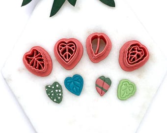 May include: A collection of pink heart and leaf-shaped polymer clay cutters. The cutters are arranged on a white surface, with green and blue leaf-shaped clay pieces. The cutters are for crafting and creating decorative elements.