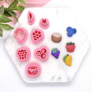 May include: A set of 12 pink plastic cookie cutters shaped like various fruits, including a strawberry, raspberry, blueberry, mango, grape, and coconut. The cutters are arranged on a white marble surface.