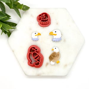 May include: Set of five red and white plastic cookie cutters shaped like bald eagles. The cutters include a full eagle, a head, and two different head profiles.