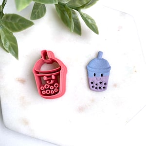 May include: Two items: a pink plastic cookie cutter and a small, hand-painted figurine. The cookie cutter is shaped like a bubble tea cup with a smiling face and boba pearls. The figurine is a blue and purple bubble tea cup with a smiling face and boba pearls.