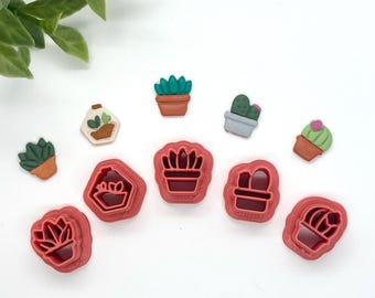 May include: A collection of cactus-themed cookie cutters and decorative succulent-shaped embellishments. The cutters are a deep red, and the succulents are various colors, including green, brown, and gray. The items are arranged on a white surface.